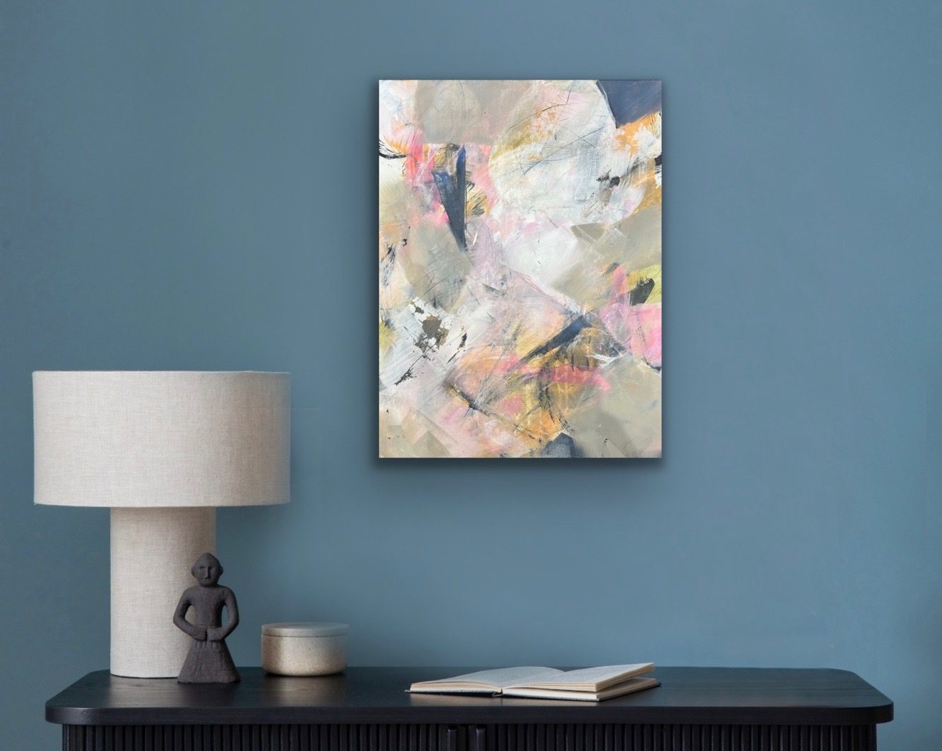 Abstract pastel-colored painting above a book and lamp on a blue wall.